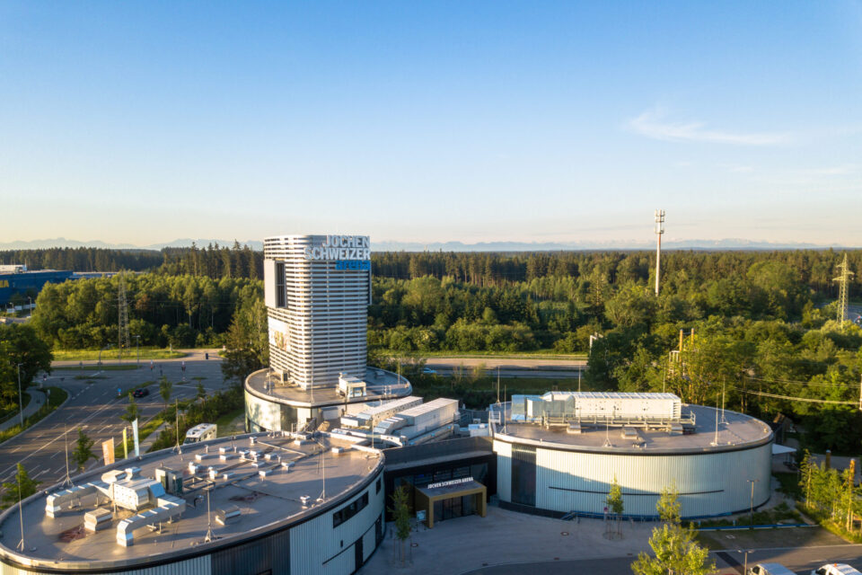 Das Bild zeigt ein modernes Gebäudekomplex inmitten von Grünflächen und Bäumen. Im Zentrum steht ein hoher, zylindrischer Turm mit einem Schriftzug am oberen Rand. Umgeben ist der Turm von flachen Gebäuden mit technischen Anlagen auf den Dächern sowie Straßen und Wegen, im Hintergrund erstreckt sich ein dichter Wald bis zum Horizont.