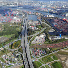 Das Bild zeigt eine Luftaufnahme eines großen Industriehafens mit zahlreichen Containern, Kränen und Frachtschiffen. Mehrere Straßen, Autobahnen und Gleise durchziehen das Gelände, das sich in der Nähe eines Gewässers befindet. Umgeben ist der Hafen von Industriegebäuden und vereinzelten Grünflächen.
