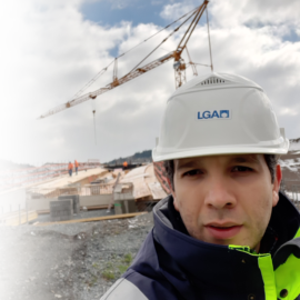 Das Bild zeigt eine Baustelle mit einem großen Kran im Hintergrund, der Baumaterialien hebt. Mehrere Arbeiter sind auf dem Gelände zu sehen, im Vordergrund steht eine Person mit weißem Schutzhelm und Warnjacke. Der Himmel ist teils bewölkt, im Hintergrund sind Hügel zu erkennen.