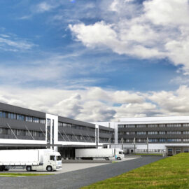 Das Bild zeigt einen großen Industrie- oder Gewerbebau mit mehreren Laderampen, an denen weiße Lastwagen geparkt sind. Das Gebäude hat mehrere Etagen mit vielen Fenstern. Im Vordergrund befindet sich eine Grasfläche, auf der zwei Personen spazieren gehen.