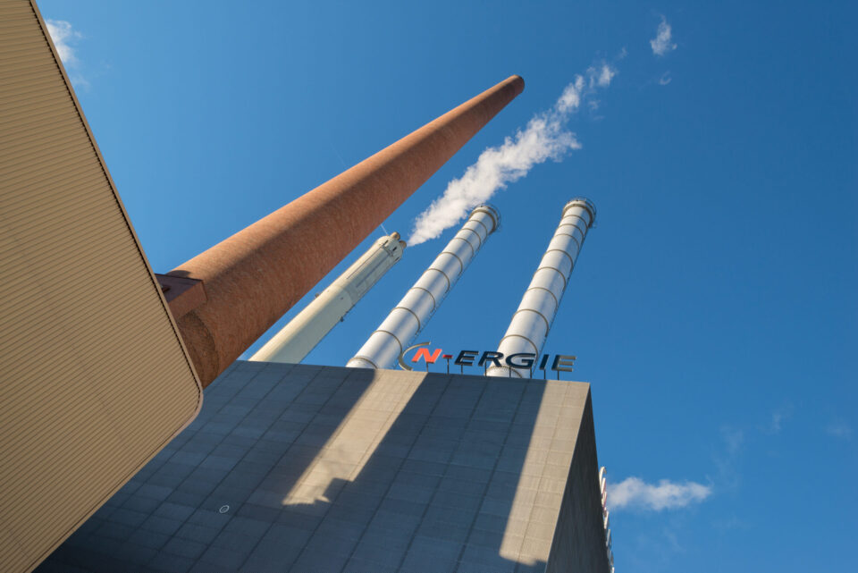 Das Bild zeigt ein Industriegebäude mit vier hohen Schornsteinen, die in den Himmel ragen. Die Schornsteine sind unterschiedlich gefärbt: rotbraun, weiß und zweimal silbern. Im Hintergrund ist ein klarer blauer Himmel mit wenigen Wolken zu sehen.