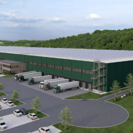 Das Bild zeigt ein großes Industrie- oder Logistikgebäude mit grüner Fassade. Vor dem Gebäude befinden sich ein Parkplatz mit vielen Autos sowie mehrere Lkw-Ladezonen. Die Anlage ist von Bäumen und Grünflächen umgeben, im Hintergrund ist ein teils bewölkter Himmel zu sehen.