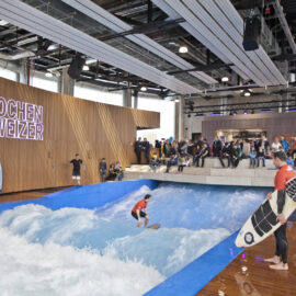 Das Bild zeigt eine Indoor-Surfanlage mit einem Wellenbecken, in dem eine Person auf einer künstlichen Welle surft. Eine weitere Person steht am Beckenrand mit einem Surfbrett in der Hand. Im Hintergrund beobachten Zuschauer das Geschehen, und an der Holzwand ist groß der Name „Jochen Schweizer“ zu sehen.