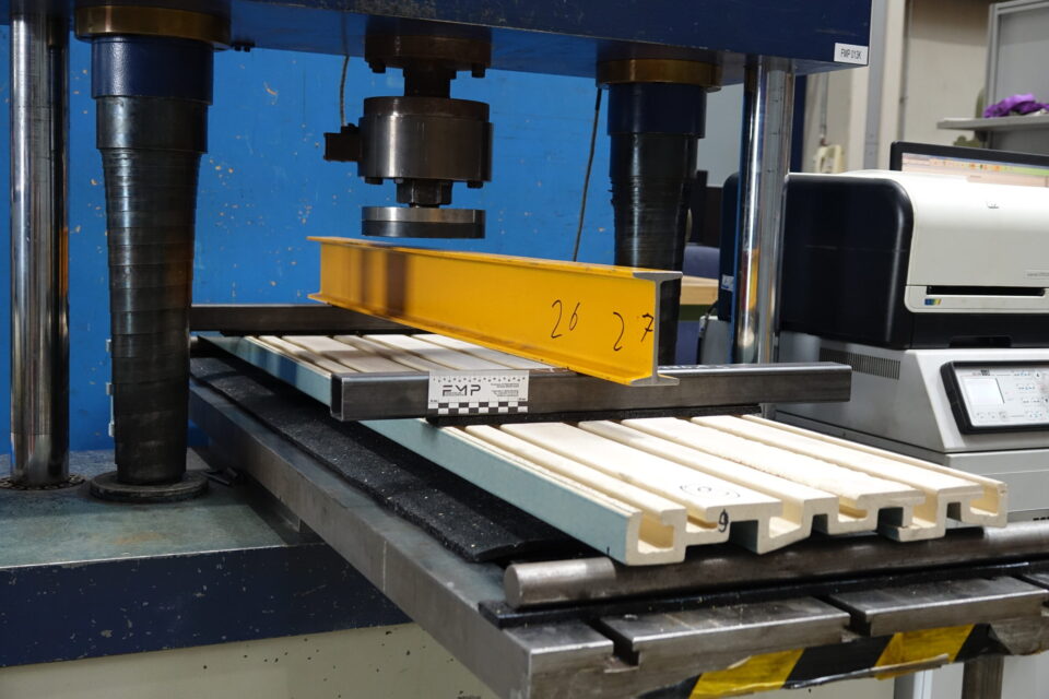 Das Bild zeigt eine große Prüfmaschine in einer Werkstattumgebung. Auf dem Tisch liegen mehrere helle Keramikleisten, die von einem gelben Stahlträger und einer metallischen Vorrichtung fixiert werden. Darüber befindet sich ein zylindrisches Prüfwerkzeug, das für Belastungstests eingesetzt wird, während im Hintergrund technische Geräte und ein blauer Maschinenrahmen sichtbar sind.