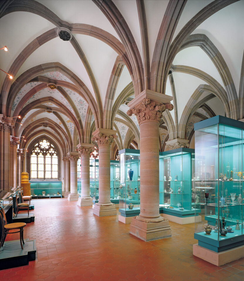 Das Bild zeigt das Innere eines Museums oder einer Galerie mit gewölbter Decke und steinernen Säulen. Am Ende des Raumes fällt Tageslicht durch große, verzierte Fenster, und mehrere Glasvitrinen präsentieren historische oder künstlerische Objekte. Der Boden ist mit roten Fliesen ausgelegt.