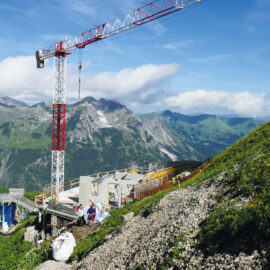 Das Bild zeigt eine Baustelle in alpiner Umgebung mit einem großen rot-weißen Kran in der Bildmitte. Unter dem Kran befinden sich mehrere im Bau befindliche Gebäude, und einige Bauarbeiter sind in der Nähe zu sehen. Im Hintergrund erstreckt sich eine Berglandschaft mit grünen Hängen und felsigen Gipfeln unter einem teils bewölkten Himmel.