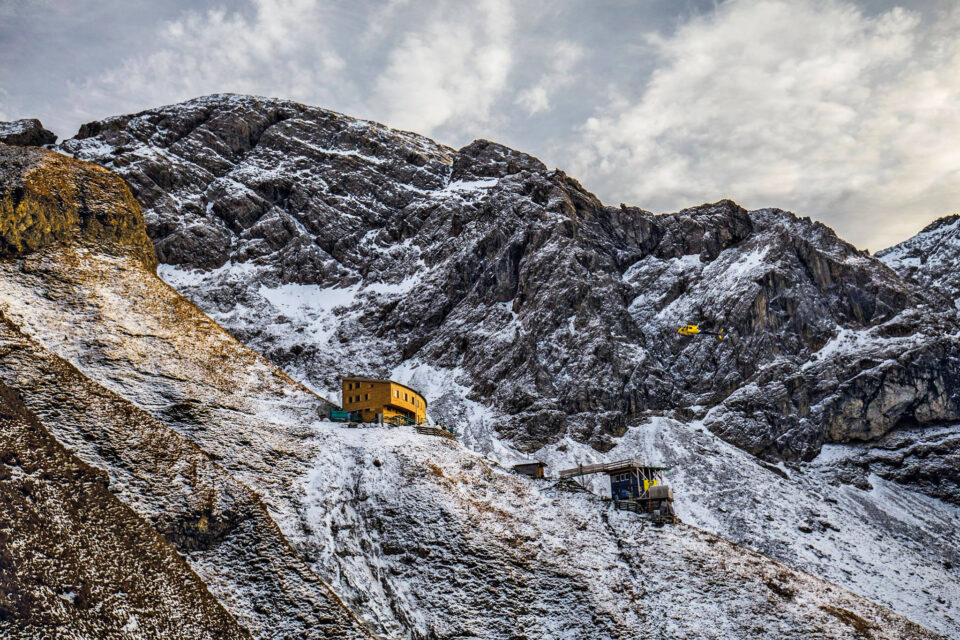 Das Bild zeigt eine verschneite Berglandschaft mit felsigen Hängen und einem Gebäude, das wie eine Berghütte aussieht. Ein gelber Hubschrauber fliegt in der Nähe des Hangs. Der Himmel ist teilweise bewölkt.