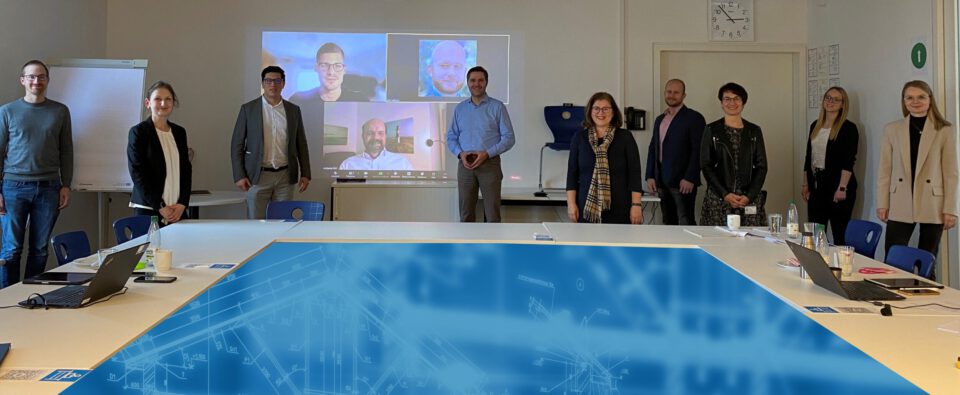 Das Bild zeigt eine Gruppe von neun Personen, die um einen Konferenztisch in einem Besprechungsraum stehen. Auf einem Bildschirm im Hintergrund sind vier weitere Teilnehmende virtuell zugeschaltet. Auf dem Tisch liegen Laptops, Dokumente, Wasserflaschen und ein technischer Plan oder eine Bauzeichnung.