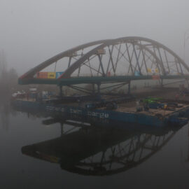 Das Bild zeigt eine große, bogenförmige Brückenkonstruktion, die auf einem Lastschiff über ein Gewässer transportiert wird. Nebel liegt über der Szene und sorgt für eine eingeschränkte Sicht, während am Ufer Baugeräte und ein Kran zu sehen sind.
