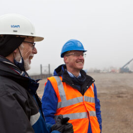 Das Bild zeigt zwei Personen mit Sicherheitshelmen und Warnkleidung auf einer Baustelle. Im Hintergrund sind Baumaschinen und Fahrzeuge zu sehen, die Szene ist in Nebel gehüllt.