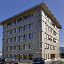Das Bild zeigt ein mehrstöckiges Gebäude mit moderner Architektur, großen Fenstern und einer Holzfassade. Vor dem Gebäude sind mehrere Autos geparkt, im Hintergrund ist etwas Grün zu sehen. Der Himmel ist klar und blau, was auf Tageslicht und gutes Wetter hinweist.