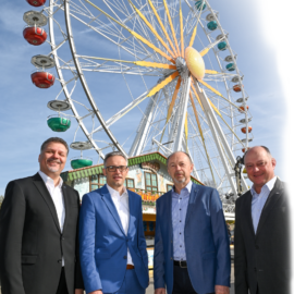Das Bild zeigt vier Personen in formeller Kleidung, die vor einem großen Riesenrad stehen. Das Riesenrad hat bunte Gondeln in Farben wie Rot, Grün, Blau und Lila. Im Hintergrund sind ein Gebäude am Fuß des Riesenrads sowie ein blauer Himmel mit einigen Wolken zu sehen.