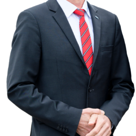 Das Bild zeigt einen Mann im Anzug mit rot-weiß gestreifter Krawatte. Der Hintergrund ist ebenfalls verschwommen, wodurch der Fokus auf der Person liegt