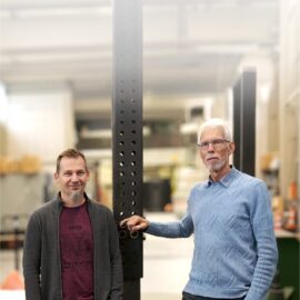 Das Bild zeigt zwei Personen, die in einer Werkhalle oder einem technischen Prüfbereich stehen. Beide lehnen an einer hohen, schwarzen Metallstütze mit zahlreichen Lochungen, die vermutlich Teil einer Prüf- oder Montagevorrichtung ist. Im Hintergrund sind Regale, Werkbänke und verschiedene Geräte zu erkennen, die auf eine industrielle oder handwerkliche Umgebung hinweisen. Die Beleuchtung ist hell und gleichmäßig, wodurch die Details der Metallkonstruktion und des Raumes gut sichtbar sind.