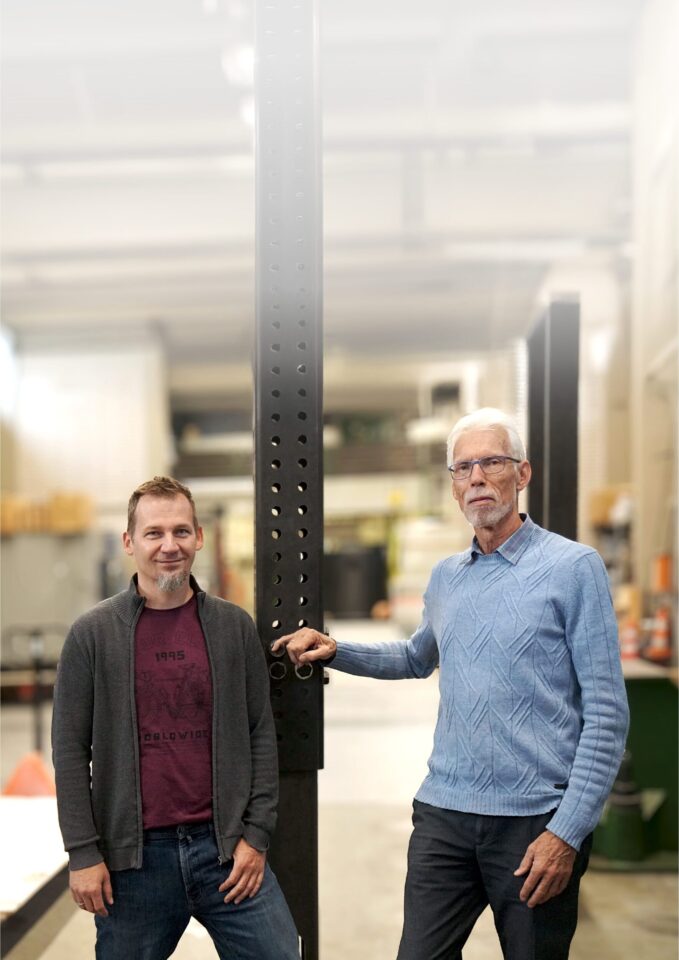 Das Bild zeigt zwei Personen, die in einer Werkhalle oder einem technischen Prüfbereich stehen. Beide lehnen an einer hohen, schwarzen Metallstütze mit zahlreichen Lochungen, die vermutlich Teil einer Prüf- oder Montagevorrichtung ist. Im Hintergrund sind Regale, Werkbänke und verschiedene Geräte zu erkennen, die auf eine industrielle oder handwerkliche Umgebung hinweisen. Die Beleuchtung ist hell und gleichmäßig, wodurch die Details der Metallkonstruktion und des Raumes gut sichtbar sind.