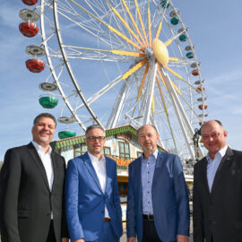 Das Bild zeigt vier Personen in formeller Kleidung, die vor einem großen Riesenrad stehen. Das Riesenrad hat bunte Gondeln in Farben wie Rot, Grün, Blau und Lila. Im Hintergrund sind ein Gebäude am Fuß des Riesenrads sowie ein blauer Himmel mit einigen Wolken zu sehen.