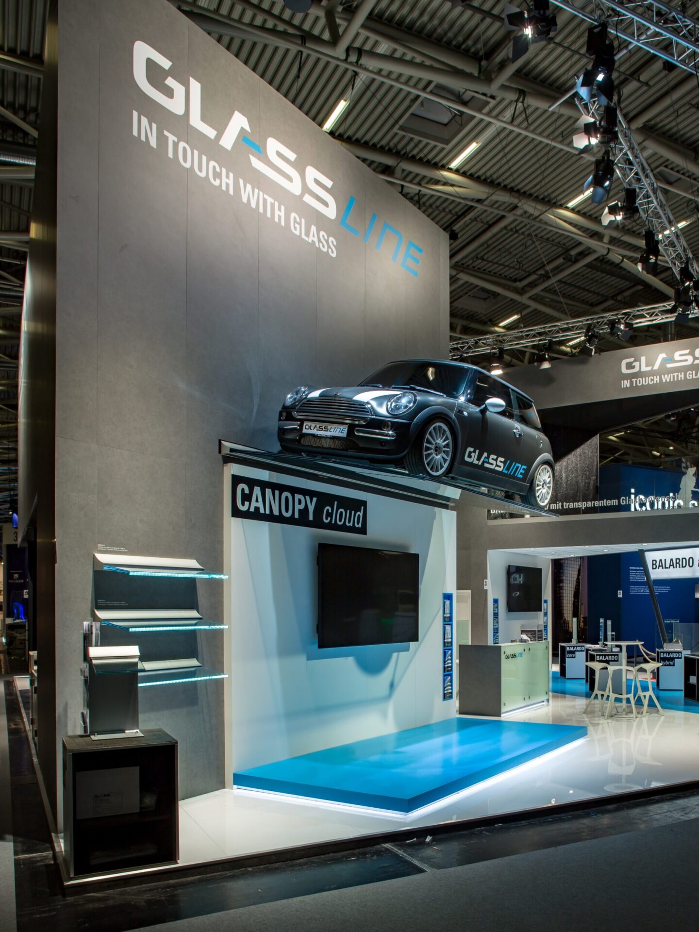 Das Bild zeigt einen modernen Messestand auf einer Fachmesse, der von hohen grauen Wänden umgeben ist. Auf der oberen Plattform des Standes steht ein schwarzes Auto, das als Blickfang dient. Unterhalb befindet sich eine Präsentationsfläche mit einem großen Bildschirm und dem Schriftzug „CANOPY cloud“. Links sind mehrere Glasplatten ausgestellt, die auf schmalen Halterungen montiert sind. Im Hintergrund sind weitere Messestände und Beleuchtungselemente der Halle sichtbar, die eine professionelle und technisch orientierte Atmosphäre schaffen.