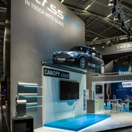 Das Bild zeigt einen modernen Messestand auf einer Fachmesse, der von hohen grauen Wänden umgeben ist. Auf der oberen Plattform des Standes steht ein schwarzes Auto, das als Blickfang dient. Unterhalb befindet sich eine Präsentationsfläche mit einem großen Bildschirm und dem Schriftzug „CANOPY cloud“. Links sind mehrere Glasplatten ausgestellt, die auf schmalen Halterungen montiert sind. Im Hintergrund sind weitere Messestände und Beleuchtungselemente der Halle sichtbar, die eine professionelle und technisch orientierte Atmosphäre schaffen.
