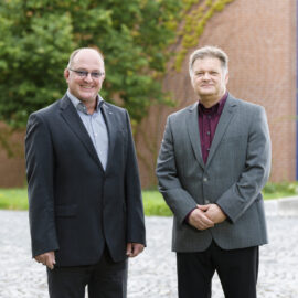 Das Bild zeigt zwei Personen im Freien auf einem gepflasterten Untergrund, im Hintergrund sind Grünflächen und ein Backsteingebäude zu sehen. Die linke Person trägt einen dunklen Anzug mit hellblauem Hemd, die rechte einen grauen Anzug mit bordeauxfarbenem Hemd.