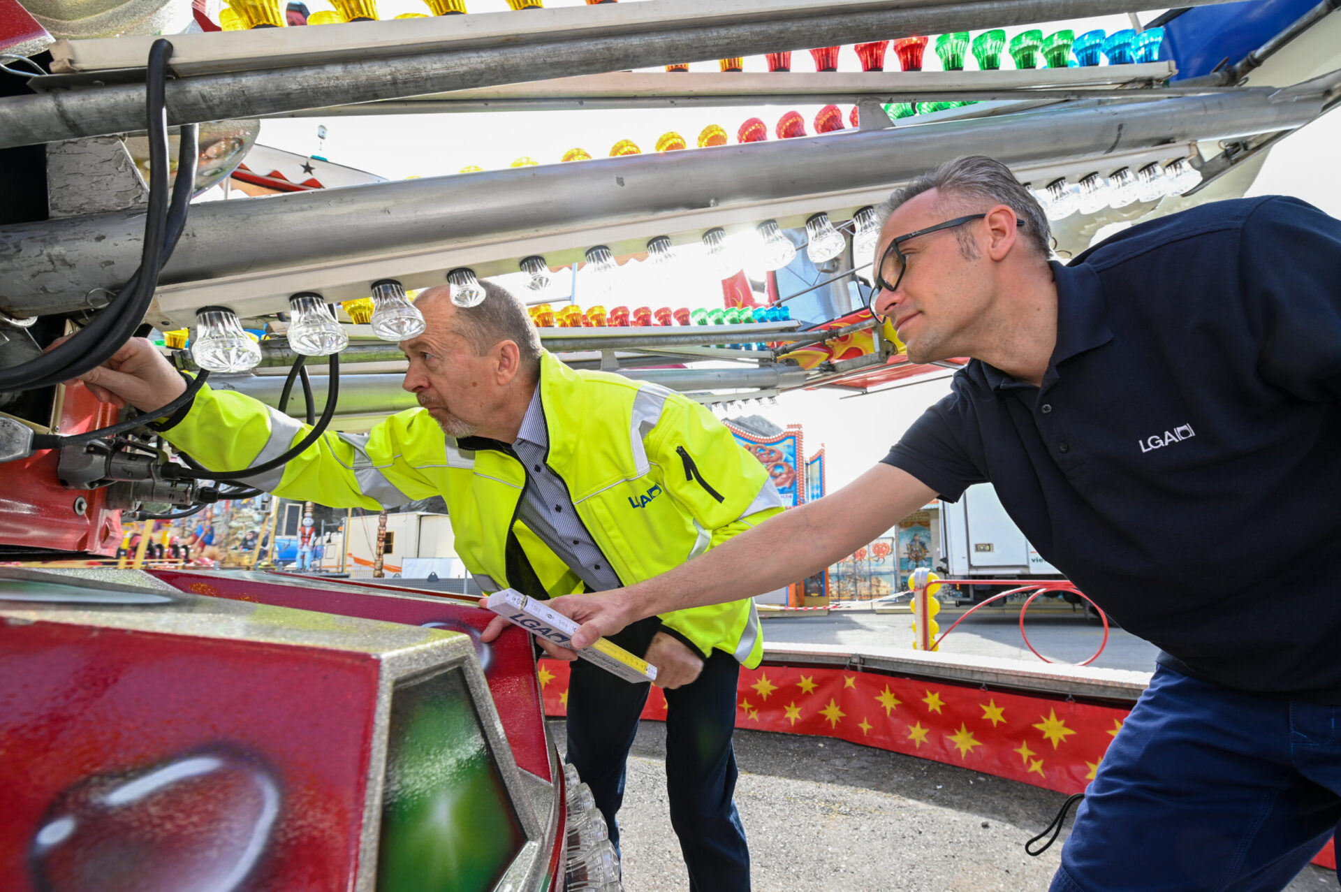 Das Bild zeigt zwei Personen, die ein Fahrgeschäft auf einem Jahrmarkt oder in einem Freizeitpark inspizieren. Eine Person trägt eine gelbe Jacke mit reflektierenden Streifen, die andere ein dunkelblaues Shirt mit Aufdruck. Sie begutachten gemeinsam die Technik des Fahrgeschäfts.