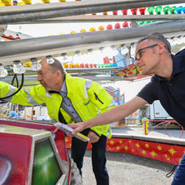 Das Bild zeigt zwei Personen, die ein Fahrgeschäft auf einem Jahrmarkt oder in einem Freizeitpark inspizieren. Eine Person trägt eine gelbe Jacke mit reflektierenden Streifen, die andere ein dunkelblaues Shirt mit Aufdruck. Sie begutachten gemeinsam die Technik des Fahrgeschäfts.