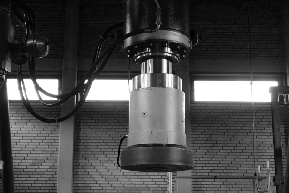 Das Bild zeigt ein großes zylindrisches Bauteil einer Industriemaschine, vermutlich Teil einer hydraulischen Presse. Mehrere Kabel und Schläuche sind an das Bauteil angeschlossen. Im Hintergrund ist eine Backsteinwand mit Fenstern zu sehen, was auf eine Werkhalle oder ein Industriegebäude hinweist.