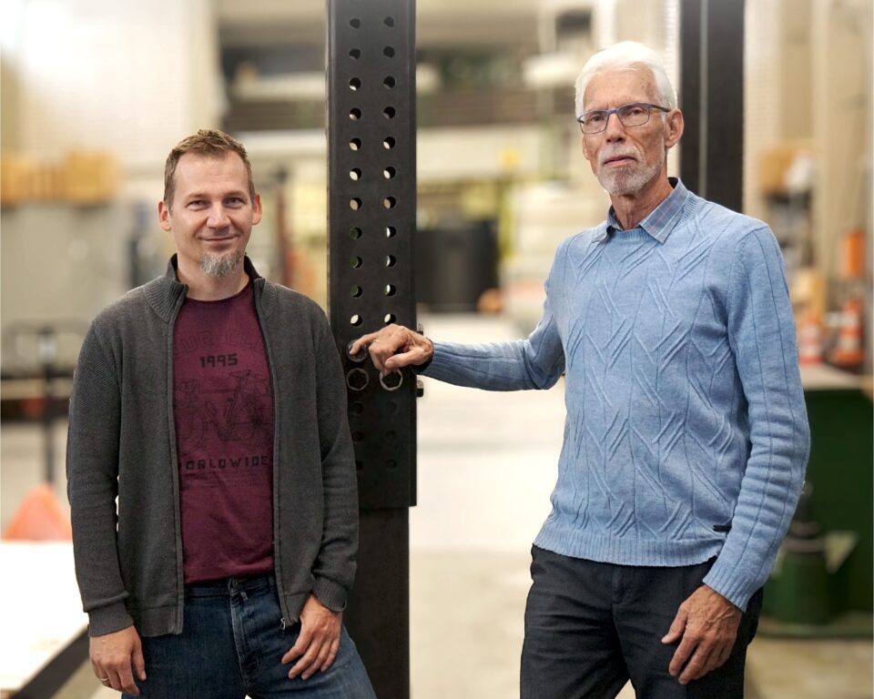 Das Bild zeigt zwei Personen, die in einer Werkhalle oder einem technischen Prüfbereich stehen. Beide lehnen an einer hohen, schwarzen Metallstütze mit zahlreichen Lochungen, die vermutlich Teil einer Prüf- oder Montagevorrichtung ist. Im Hintergrund sind Regale, Werkbänke und verschiedene Geräte zu erkennen, die auf eine industrielle oder handwerkliche Umgebung hinweisen. Die Beleuchtung ist hell und gleichmäßig, wodurch die Details der Metallkonstruktion und des Raumes gut sichtbar sind.