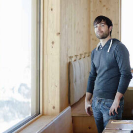 Das Bild zeigt eine Person in einem Raum mit Holzverkleidung, die aus einem großen Fenster blickt. Die Person trägt einen grauen Hoodie und Jeans. Entlang der Wand befinden sich gepolsterte Sitzgelegenheiten aus Holz.