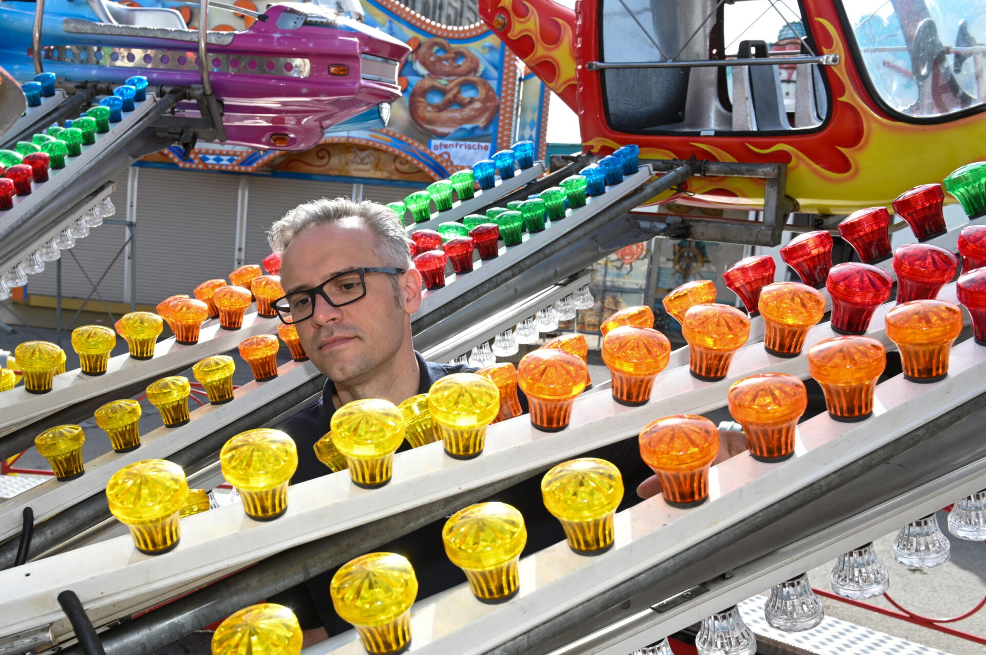 Das Bild zeigt eine Person, die hinter mehreren Reihen bunter Glühbirnen steht, die auf schrägen Metallschienen angeordnet sind. Die Glühbirnen leuchten in Farben wie Gelb, Orange, Rot und Grün. Im Hintergrund ist ein Teil eines Fahrgeschäfts mit einem lilafarbenen Wagen und weiteren bunten Dekorationen zu sehen.