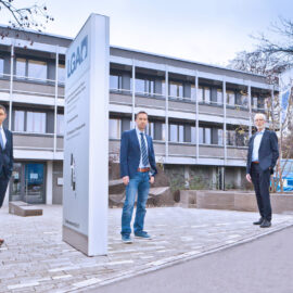 Das Bild zeigt drei Personen in Businesskleidung vor einem Gebäude mit der Aufschrift „LGA“. Im Hintergrund steht ein Auto mit dem LGA-Logo, daneben befinden sich Fahnen mit dem gleichen Logo.