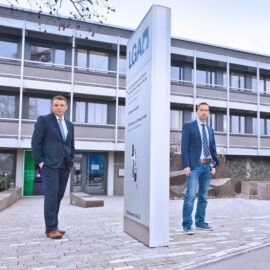 Das Bild zeigt zwei Personen in Businesskleidung vor einem Gebäude mit mehreren Fenstern. Im Vordergrund steht ein Schild mit der Aufschrift „LGA“ und weiteren, schwer lesbaren Textzeilen. Das Gebäude wirkt wie ein Büro- oder Institutsgebäude.