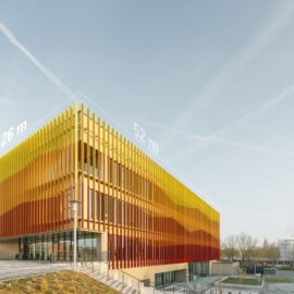 Das Bild zeigt ein modernes Gebäude mit einer auffälligen Fassadengestaltung aus vertikalen Lamellen in Gelb-, Orange- und Rottönen, die einen sanften Farbverlauf bilden. Die Lamellen sind vor einer Glasfassade angebracht und verleihen dem Bauwerk ein markantes, dynamisches Erscheinungsbild. Auf dem Dach sind Maßangaben von 26 Metern Breite und 52 Metern Länge eingeblendet, während im Hintergrund ein klarer Himmel mit Kondensstreifen sowie einige Industrieanlagen zu sehen sind.