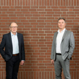 Das Bild zeigt zwei Personen in formeller Kleidung, die vor einer Ziegelwand stehen. Die linke Person trägt einen dunklen Anzug mit hellblauem Hemd, die rechte einen hellgrauen Anzug mit weißem Hemd.