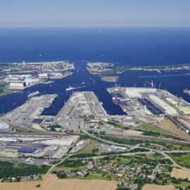 Das Bild zeigt eine Luftaufnahme eines großen Hafengebiets mit mehreren Anlegestellen, Lagerhallen und Industriegebäuden entlang eines Flusses, der ins Meer mündet. Mehrere Schiffe liegen an den Kais, und Kräne sind in den Industrieabschnitten zu erkennen. Die Umgebung umfasst städtische Bereiche, Grünflächen und landwirtschaftlich genutzte Felder, im Hintergrund ist eine weite Wasserfläche mit vielen kleinen Booten zu sehen.