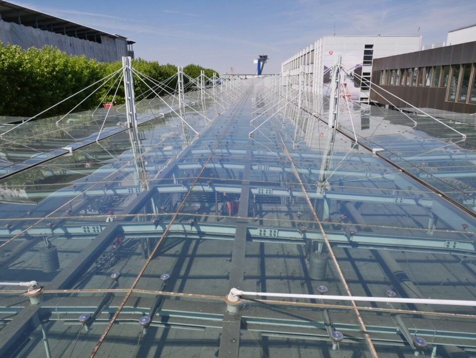 Das Bild zeigt eine große Glasdachkonstruktion mit Metallträgern und Seilverspannungen. Links ist etwas Begrünung zu sehen, rechts moderne Gebäude, die zu einem gewerblichen oder industriellen Komplex gehören. Der Himmel ist klar und sonnig.