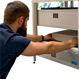 Das Bild zeigt eine Person mit Bart und kurzem Haar, die ein Objekt in ein technisches Gerät einsetzt oder justiert. Das Gerät wirkt wie ein Labor- oder Industrieapparat mit mehreren Fächern und Bedienelementen.