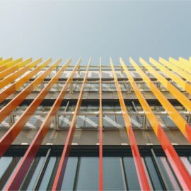 Das Bild zeigt eine Nahaufnahme der Fassade eines modernen Gebäudes, aufgenommen aus einer Perspektive von unten nach oben. Auffällig sind die vertikalen Lamellen in kräftigen Gelb-, Orange- und Rottönen, die vor einer Glasfassade angebracht sind und einen dynamischen Farbverlauf bilden. Hinter den Lamellen sind Metallgitter und Teile der Fensterstruktur sichtbar, während der Hintergrund einen klaren Himmel zeigt.