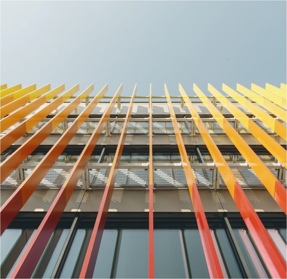 Das Bild zeigt eine Nahaufnahme der Fassade eines modernen Gebäudes, aufgenommen aus einer Perspektive von unten nach oben. Auffällig sind die vertikalen Lamellen in kräftigen Gelb-, Orange- und Rottönen, die vor einer Glasfassade angebracht sind und einen dynamischen Farbverlauf bilden. Hinter den Lamellen sind Metallgitter und Teile der Fensterstruktur sichtbar, während der Hintergrund einen klaren Himmel zeigt.