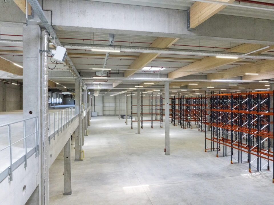 Das Bild zeigt einen Weg aus hellen Pflastersteinen entlang einer Gebäudewand aus Metall. Links am Weg wachsen junge Weinreben an Drahtgestellen, eine davon ist mit einem Schild mit der Aufschrift „Weintraube“ und einem Traubenbild versehen. Rechts des Weges befindet sich ein trockener Hang mit spärlicher Vegetation und einer Betonmauer am oberen Ende.