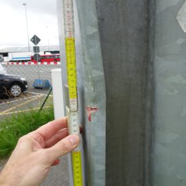 Das Bild zeigt eine Hand, die ein Maßband an eine Metalloberfläche hält, wobei das Band auf etwa 27 Zentimeter ausgezogen ist. In der Nähe der 22-Zentimeter-Marke befindet sich eine kleine rote Markierung auf dem Metall. Im Hintergrund sind Fahrzeuge wie ein schwarzes Auto und ein roter Bus sowie Straßenschilder und Absperrungen zu sehen.