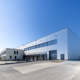 Das Bild zeigt ein großes Industrie- oder Lagergebäude mit moderner weißer Fassade, mehreren Laderampen und großen Fenstern im oberen Bereich. Vor dem Gebäude befindet sich eine weitläufige asphaltierte Fläche für Fahrzeuge. Der Himmel ist klar und blau.