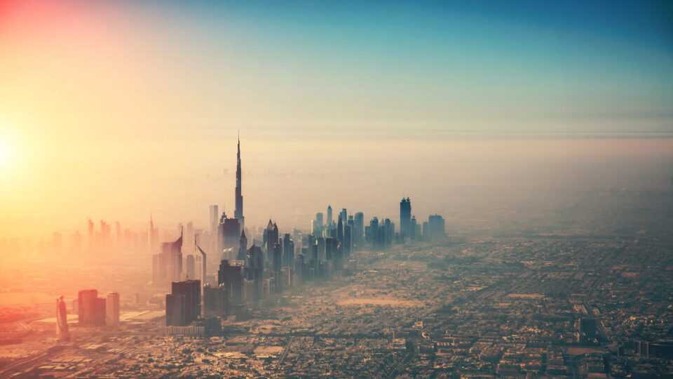 Das Bild zeigt eine Luftaufnahme einer Stadt mit zahlreichen Hochhäusern, im Zentrum ragt der Burj Khalifa als höchstes Gebäude hervor. Die Sonne geht links am Horizont auf oder unter und taucht die Stadt in ein warmes Licht. Der Himmel ist klar mit einem Farbverlauf von Blau zu Orange.