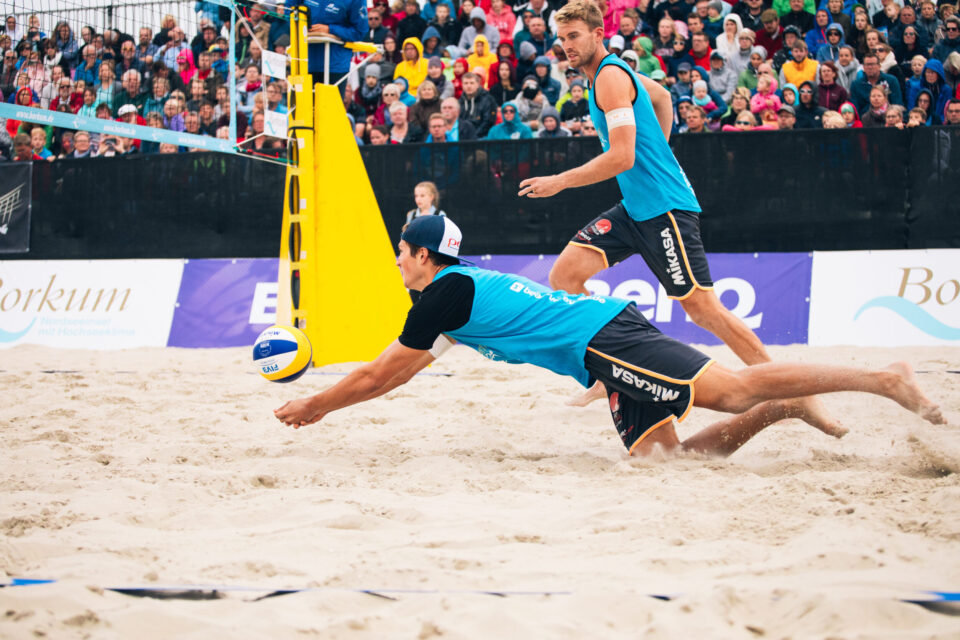 Das Bild zeigt ein Beachvolleyballspiel mit zwei Spielern im Vordergrund, beide in blauen Trikots und schwarzen Shorts. Einer der Spieler hechtet mit ausgestrecktem Arm nach dem Ball, während der andere im Hintergrund steht und bereit ist zu reagieren. Im Hintergrund sind ein gelber Netzpfosten, Zuschauertribünen und Werbebanner zu sehen.