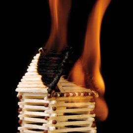 Das Bild zeigt ein kleines Hausmodell aus Streichhölzern, das in Flammen steht. Die Feuerzungen erfassen vor allem den oberen Bereich des Hauses, wobei die Streichhölzer sichtbar verkohlen. Der schwarze Hintergrund hebt die orange-gelben Flammen deutlich hervor.