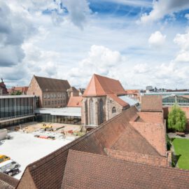 Das Bild zeigt eine historische Gebäudeanlage mit roten Ziegeldächern aus der Vogelperspektive. In der Mitte befindet sich ein Innenhof mit grünem Rasen und Bäumen, umgeben von modernen Glasbauten. Der Himmel ist teils bewölkt mit blauen Abschnitten.