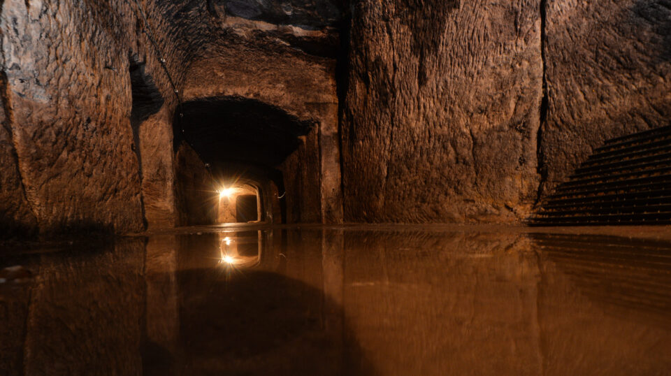 Das Bild zeigt einen unterirdischen Tunnel mit gemauerten Steinwänden und einem gewölbten Deckengewölbe. Auf dem Boden befindet sich eine Wasserfläche, die das Licht aus einer entfernten Lichtquelle im Tunnel reflektiert.