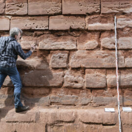 Das Bild zeigt eine Person, die eine große Mauer aus rechteckigen Steinblöcken erklimmt oder untersucht. Die Person trägt ein kariertes Hemd, Jeans und eine Umhängetasche. Rechts neben ihr steht ein senkrecht angelegter Messstab, am Boden befinden sich zwei kleine weiße Tafeln und ein Werkzeug.