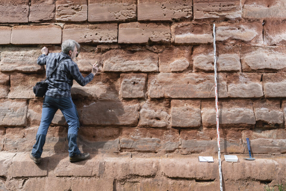 Das Bild zeigt eine Person, die eine große Mauer aus rechteckigen Steinblöcken erklimmt oder untersucht. Die Person trägt ein kariertes Hemd, Jeans und eine Umhängetasche. Rechts neben ihr steht ein senkrecht angelegter Messstab, am Boden befinden sich zwei kleine weiße Tafeln und ein Werkzeug.
