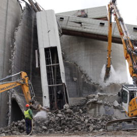 Auf dem Bild ist eine Abbruchstelle zu sehen, auf der zwei Bagger ein großes Betonbauwerk niederreißen. Ein Arbeiter mit grünem Schutzanzug und rotem Helm sprüht Wasser auf die Trümmer, um Staub zu binden. Die Szene zeigt eine fortgeschrittene Phase des Rückbaus mit deutlich sichtbaren Schäden am Bauwerk.