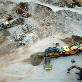 Das Bild zeigt eine Luftaufnahme eines Steinbruchs oder Bergbaugeländes. Zwei große Maschinen, vermutlich Brecher oder Siebanlagen, verarbeiten das geförderte Material. Jeweils ein orangefarbener Bagger belädt die Maschinen, daneben stehen gelbe Container mit verarbeitetem Material.